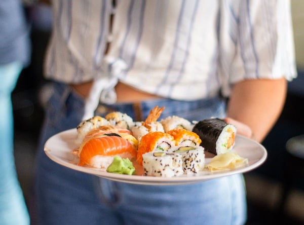 Eine Frau im gestreiftem Hemd und blauer Jeans hält einen Teller mit verschiedenen Sushi Arten